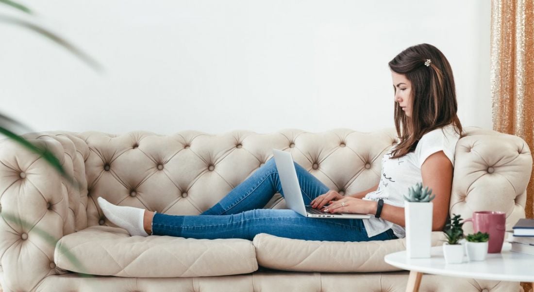 A casually dressed young woman working from home on a laptop, stretched out on a sofa next to a coffee table with a mug and some small potted plants.