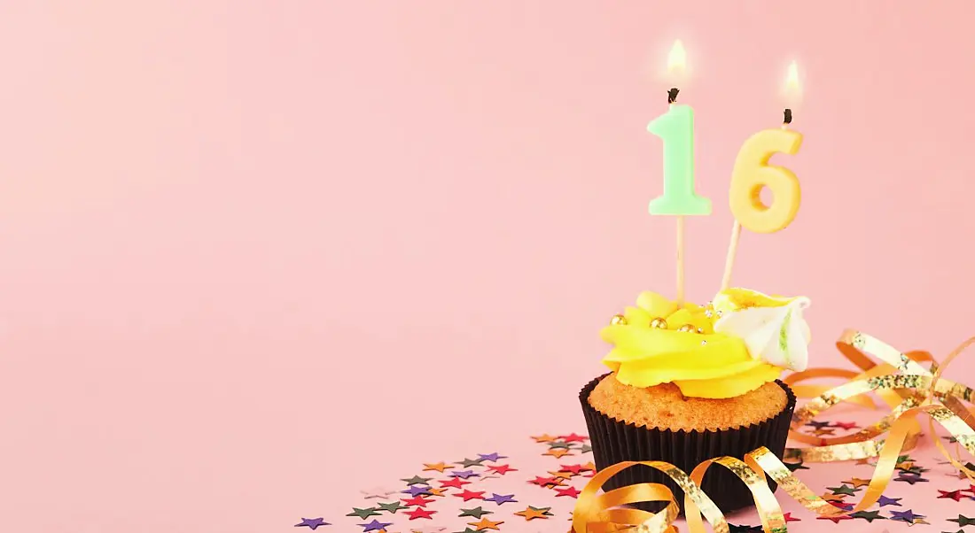 A cupcake with a 16 candle is sitting on a table against a pink background.