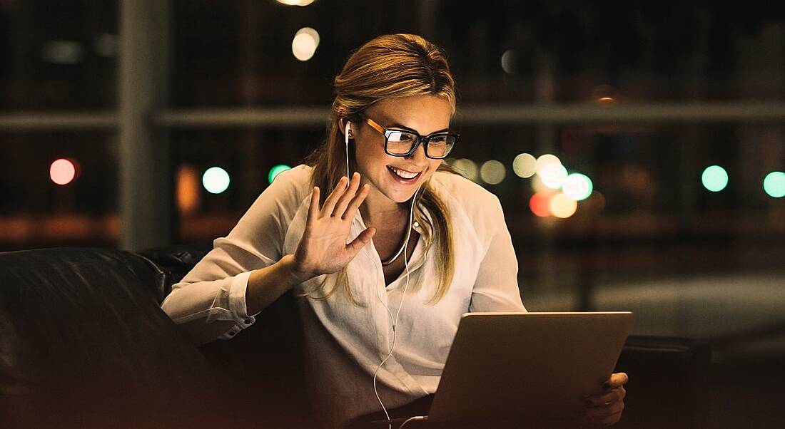 Woman wearing glasses and a white top waving to a laptop screen in a dimly lit room at night.