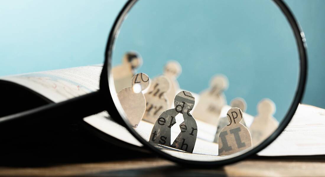 Recruitment or hiring concept with icons of candidates made from paper standing on an open newspaper under a magnifying glass.