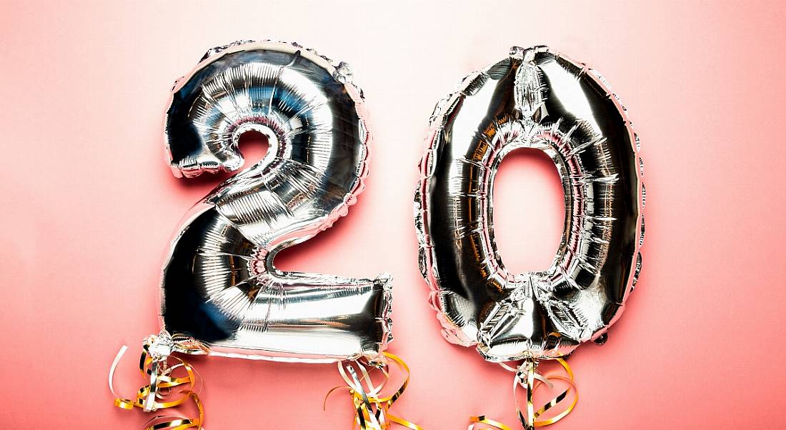 Two inflated silver balloons with curling strings attached on a soft pink background. The balloons read ‘20’.