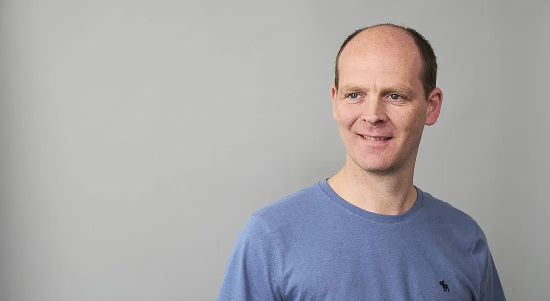 Dave Anderson is wearing a blue T-shirt and standing against a pale backdrop.