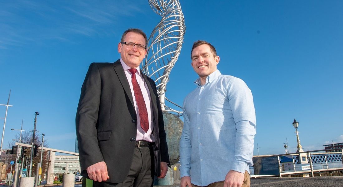 Derek Andrews in a black suit with red tie, and Conor O’Loughlin in a blue shirt and grey chinos, standing in front of a wire statue against a sunny sky.