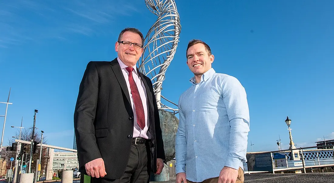 Derek Andrews in a black suit with red tie, and Conor O’Loughlin in a blue shirt and grey chinos, standing in front of a wire statue against a sunny sky.