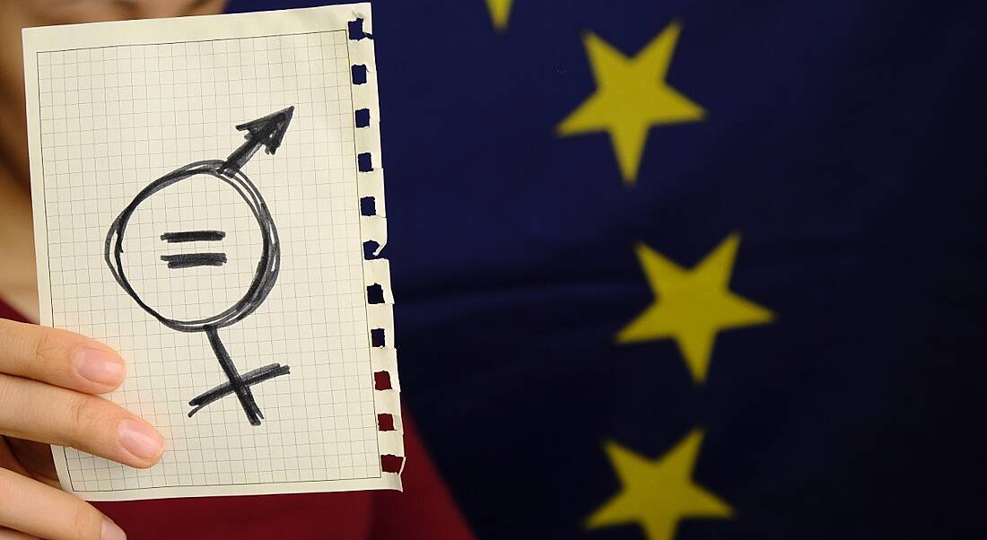 Person holding up a sheet of paper with symbols for gender equality on it, with the EU flag in the background.