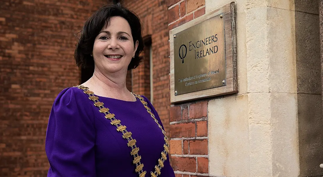 Marguerite Sayers is standing outside of the Engineers Ireland building and wearing a purple dress while smiling into the camera.