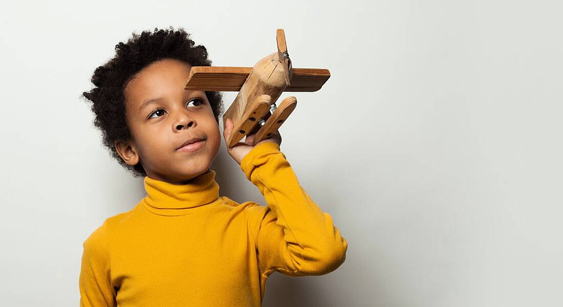 A boy in a yellow jumper playing with a wooden plane model.