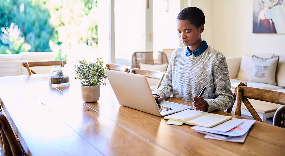 A woman is using a laptop and writing notes while working from home, symbolising managing a team working remotely.