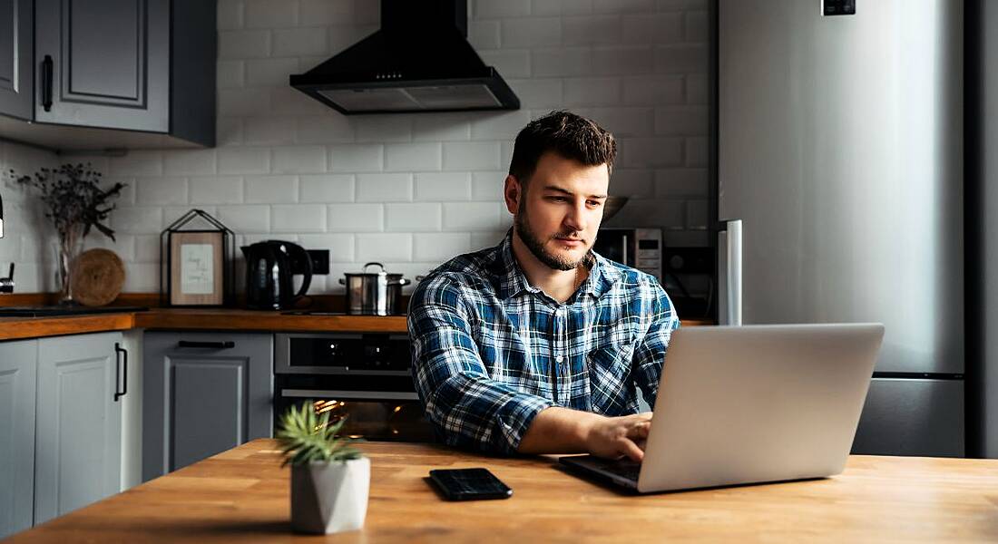 A man with a laptop sits at his kitchen table. You can see kitchen counters and a hob behind him.