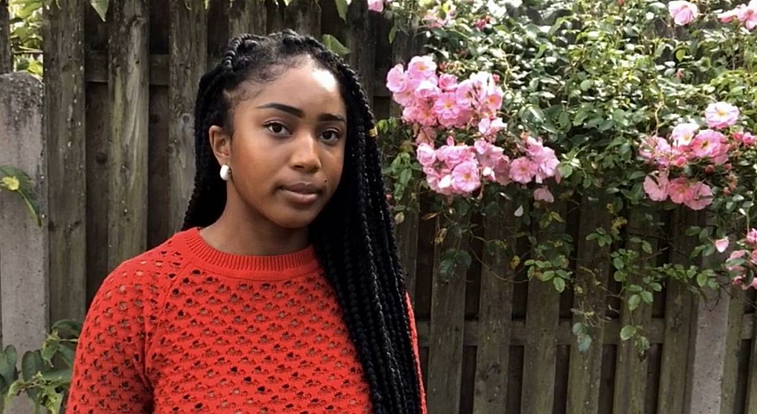 Computer science graduate Oluchi Anyabuike is standing outside and smiling into the camera while wearing a red jumper and standing beside pink flowers.