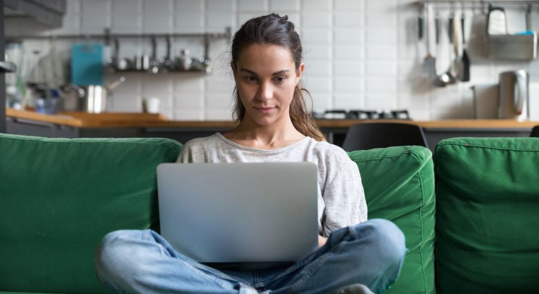 A woman is sitting on a green couch and working on a laptop in her home.