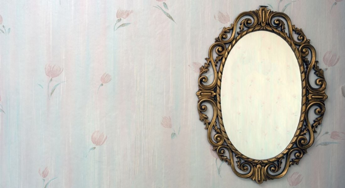 An old-fashioned mirror hanging on a wall with floral wallpaper, symbolising beauty and lookism.