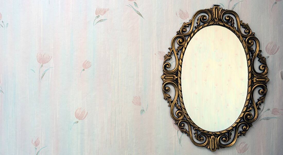 An old-fashioned mirror hanging on a wall with floral wallpaper, symbolising beauty and lookism.