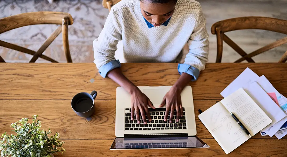 Top-down view of a woman working from home on a wooden desk with a laptop, plant and stationery.