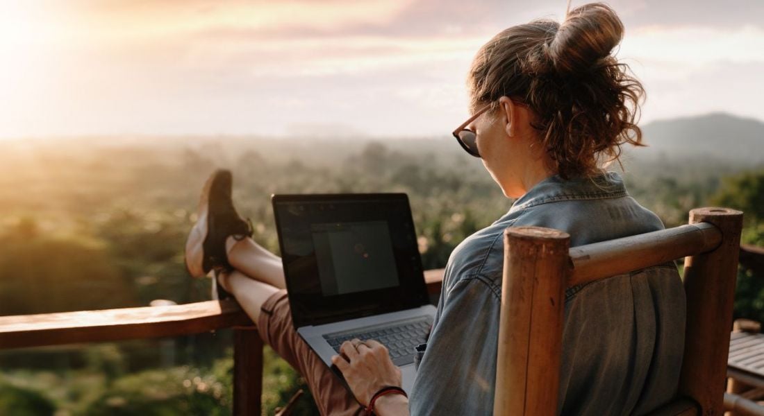 A woman works on her laptop with her feet up on the rail of a balcony overlooking a sunrise over a forest.