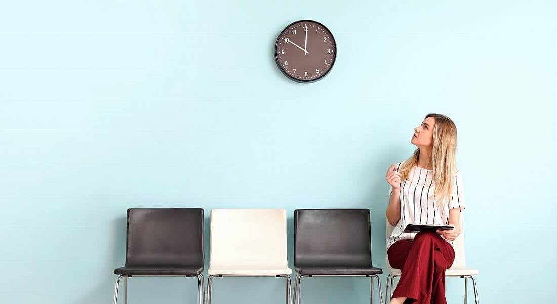 A woman is sitting down on a row of chairs against a blue wall, looking up anxiously at a clock on the wall.