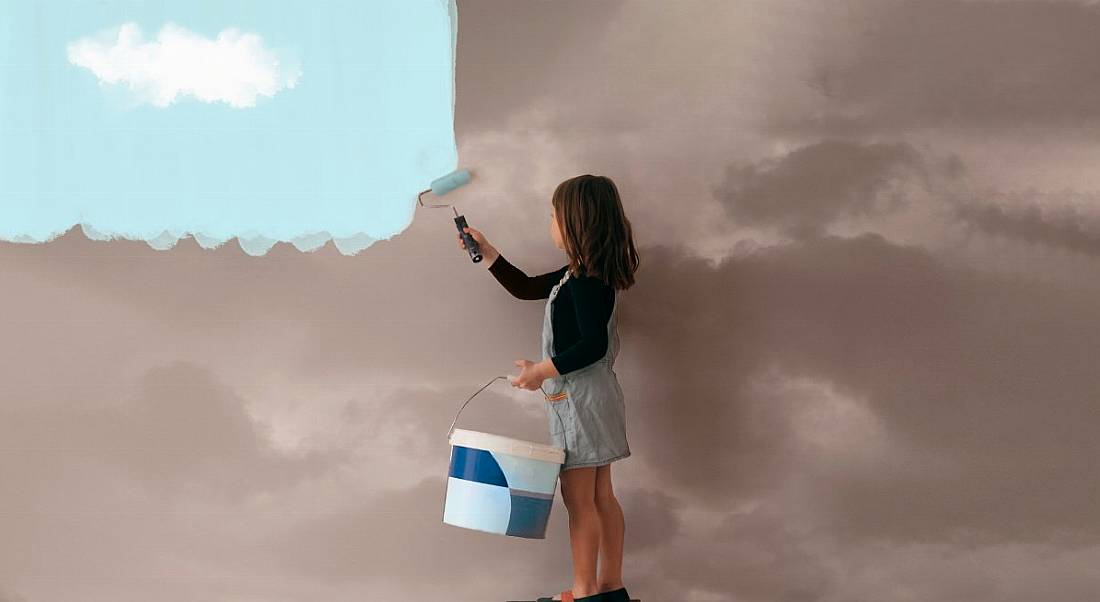 Little girl uses a can of paint to colour the wall of a room from cloudy grey to clear blue sky.