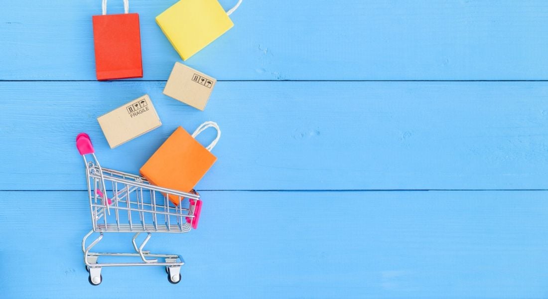 Colourful shopping bags are falling into a shopping trolley against a blue background, symbolising e-commerce and online shopping sites such as eShopWorld.