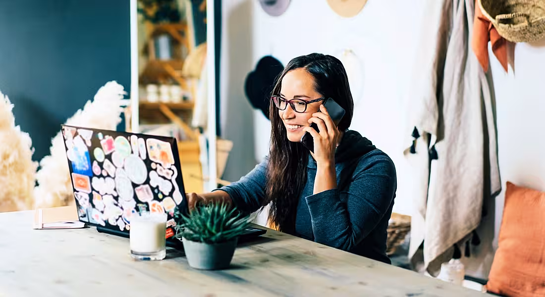 A woman wearing glasses is working remotely while talking on the phone and smiling.