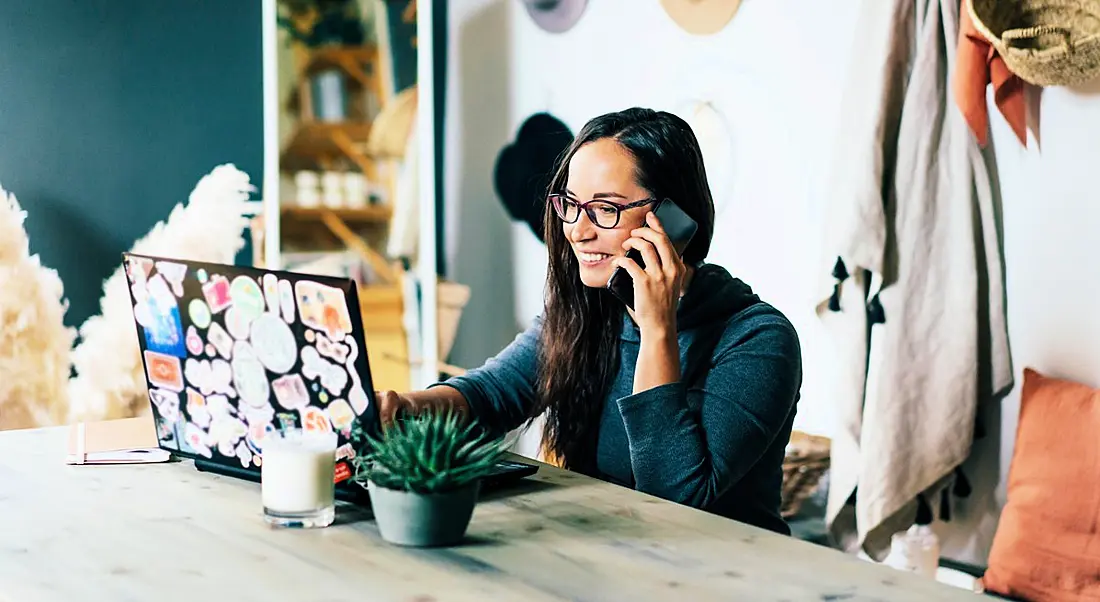 A woman wearing glasses is working remotely while talking on the phone and smiling.