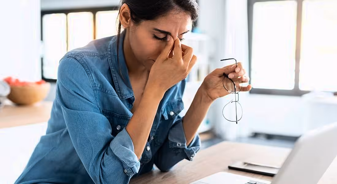 A stressed woman is working from home, pinching the bridge of her nose while sitting at a laptop.