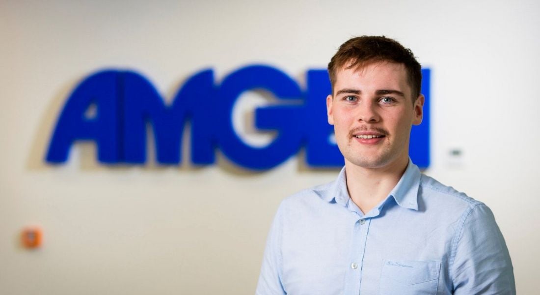 Electrical-engineering graduate Fintan Naughton is standing against a wall with a blue Amgen sign.