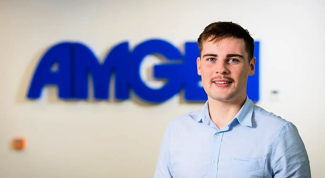 Electrical-engineering graduate Fintan Naughton is standing against a wall with a blue Amgen sign.