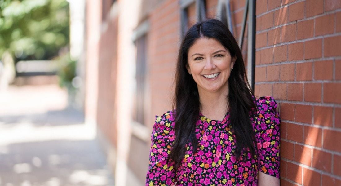 Katie Burke of HubSpot is standing outside in a pink dress, smiling into the camera while leaning agaisnt a red-brick wall.