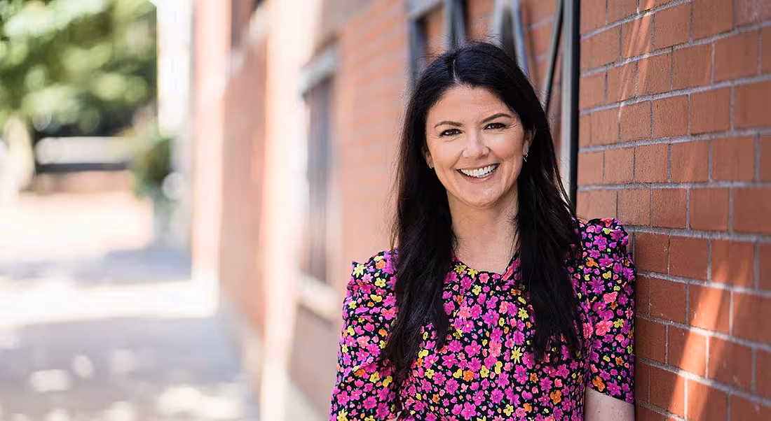 Katie Burke of HubSpot is standing outside in a pink dress, smiling into the camera while leaning agaisnt a red-brick wall.