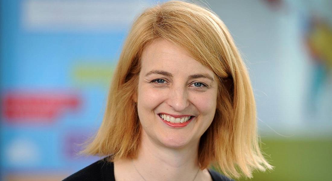 A headshot of a woman with short red hair, smiling at the camera. She is standing in front of an out-of-focus colourful background.