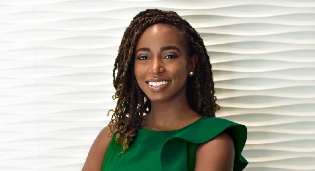 Brittany S Hale of BND Consulting is wearing a green dress and standing against an off-white wall, smiling into the camera with her arms folded.