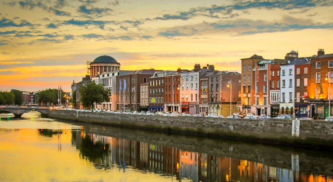 A view of the quays in Dublin city. The River Liffey is in the foreground and a long line of buildings is in the background against an orange sunset sky.
