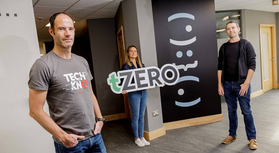 Senior employees at Overstock are standing in an office setting and smiling into the camera. One is holding up a tZero sign.