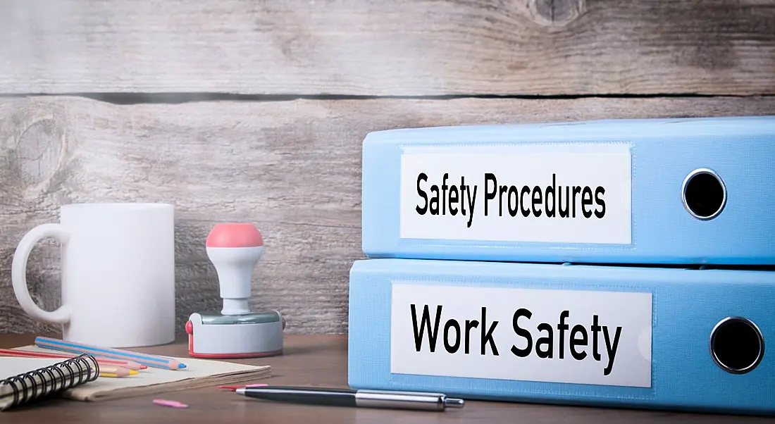 Two binders are sitting on a desk beside a cup of coffee. One binder has a label that reads 'Work Safety', the other says 'Safety Procedures'.