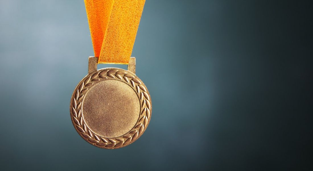 A gold medal with a yellow strap is handing against a dark green background.