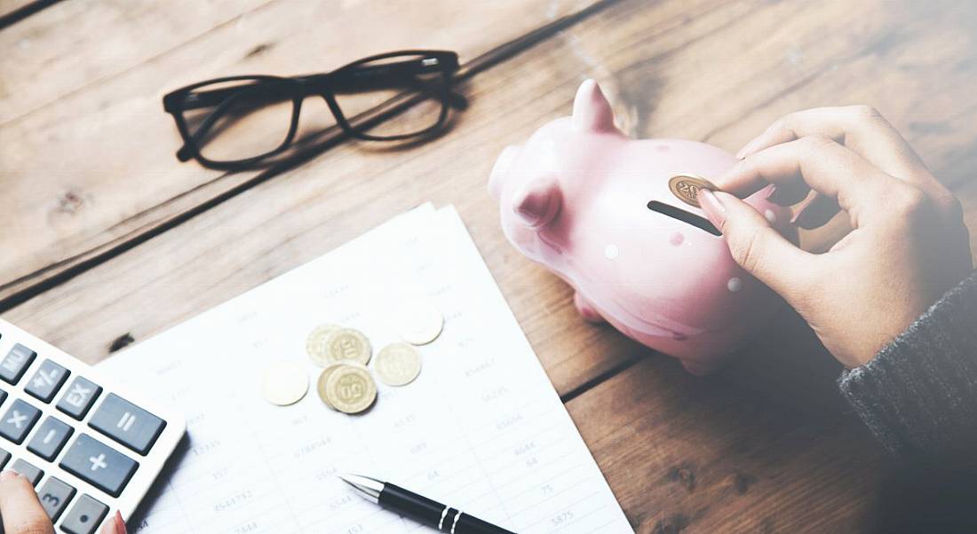 A person is putting a coin into a piggy bank beside glasses and a calculator on a wooden desk.