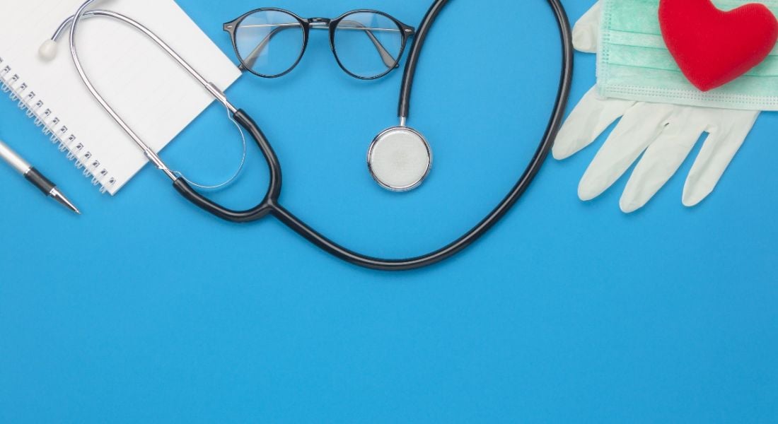 A stethoscope, face mask, toy heart, glasses and a notebook are laying against a blue background, symbolising future health trends and the future of work.