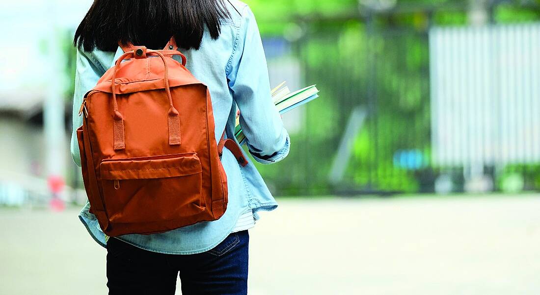 A student is facing away from the camera wearing an orange backpack and carrying books in an outdoor setting.