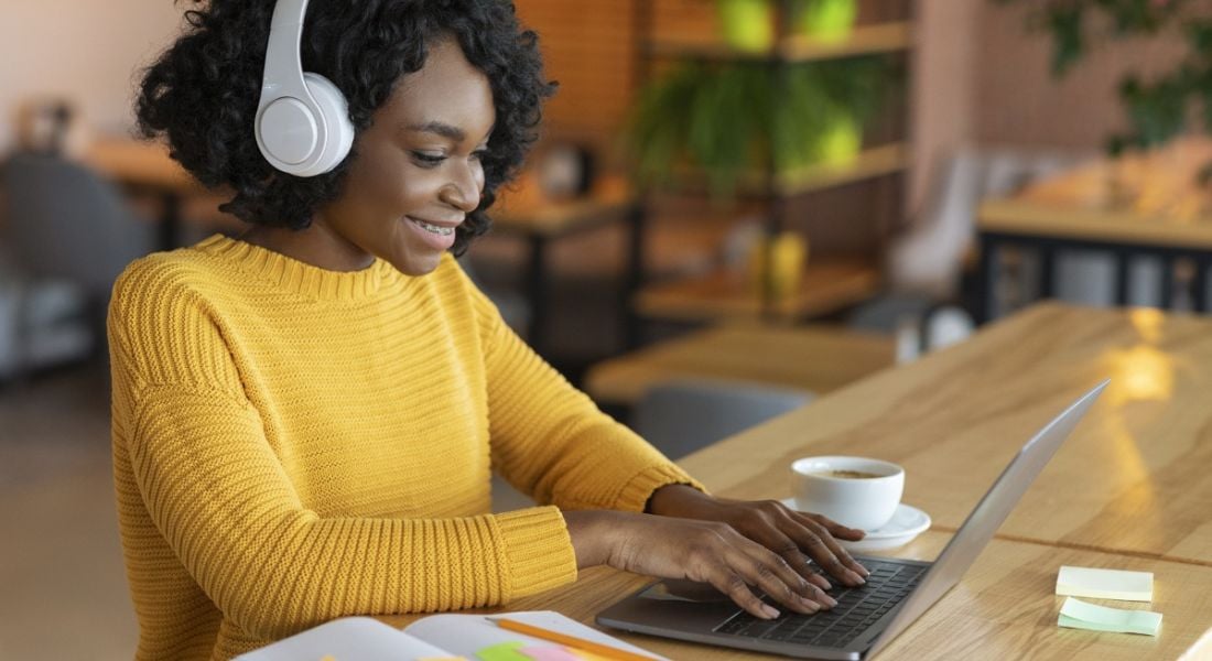 A professional woman in a bright yellow jumper is sitting and working on a laptop in a café.