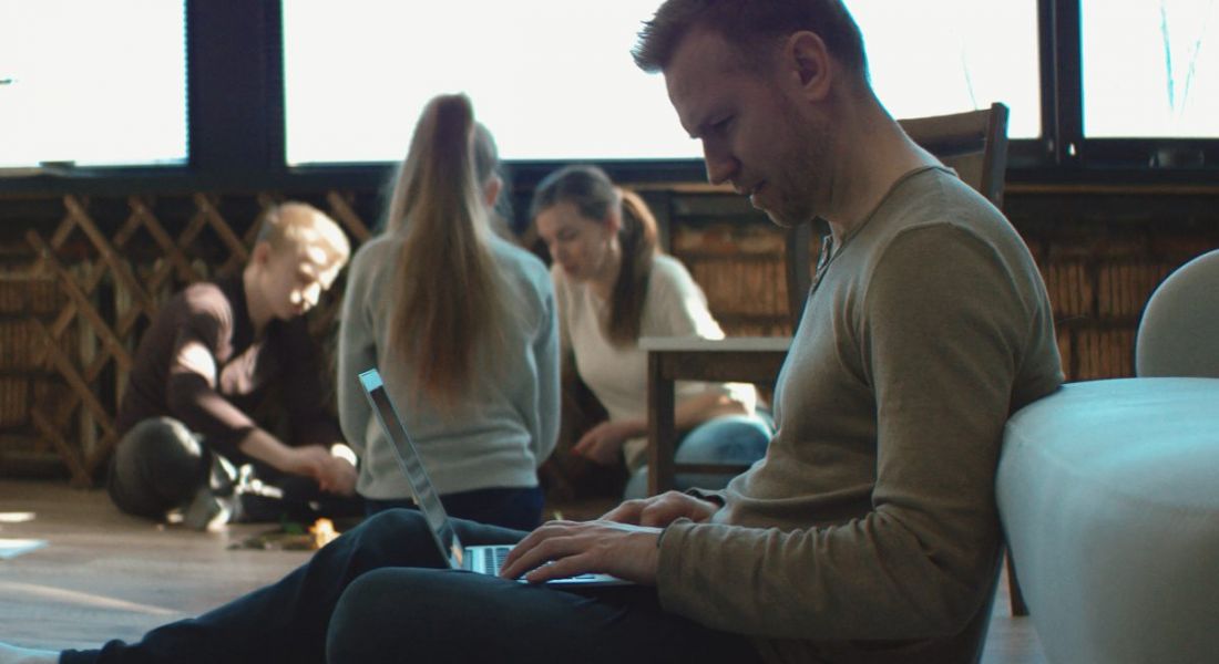 A man is sitting on the floor at home working on his laptop remotely while his children play in the background.