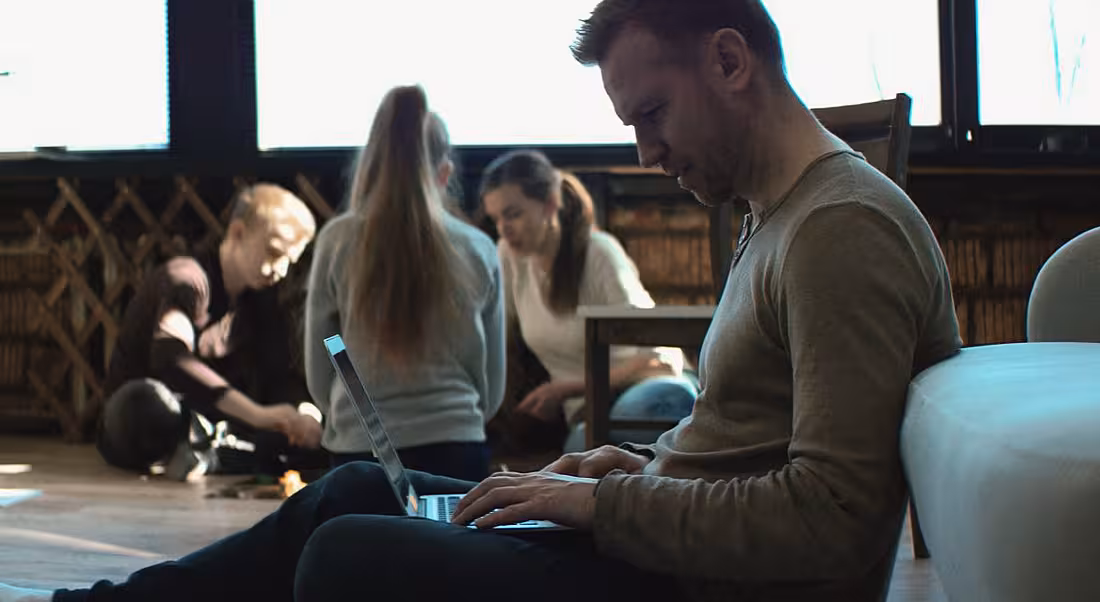 A man is sitting on the floor at home working on his laptop remotely while his children play in the background.