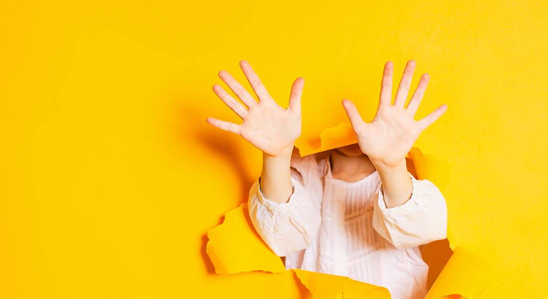 A girl is pushing through a yellow background and holding her 10 fingers up.