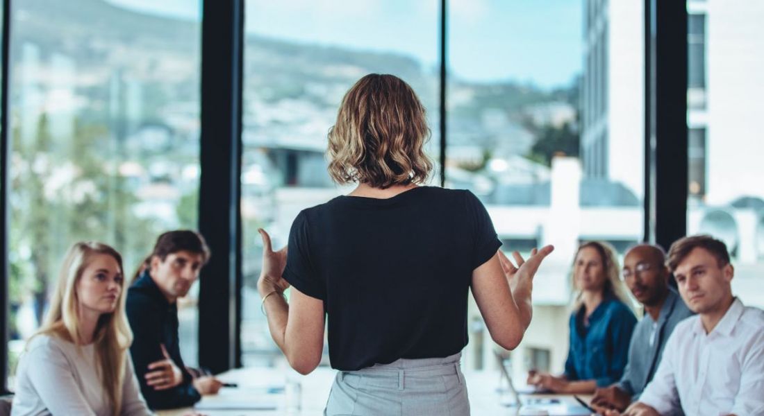 A woman leader is standing up and speaking to a team of colleagues who are sitting down in a brightly lit office.