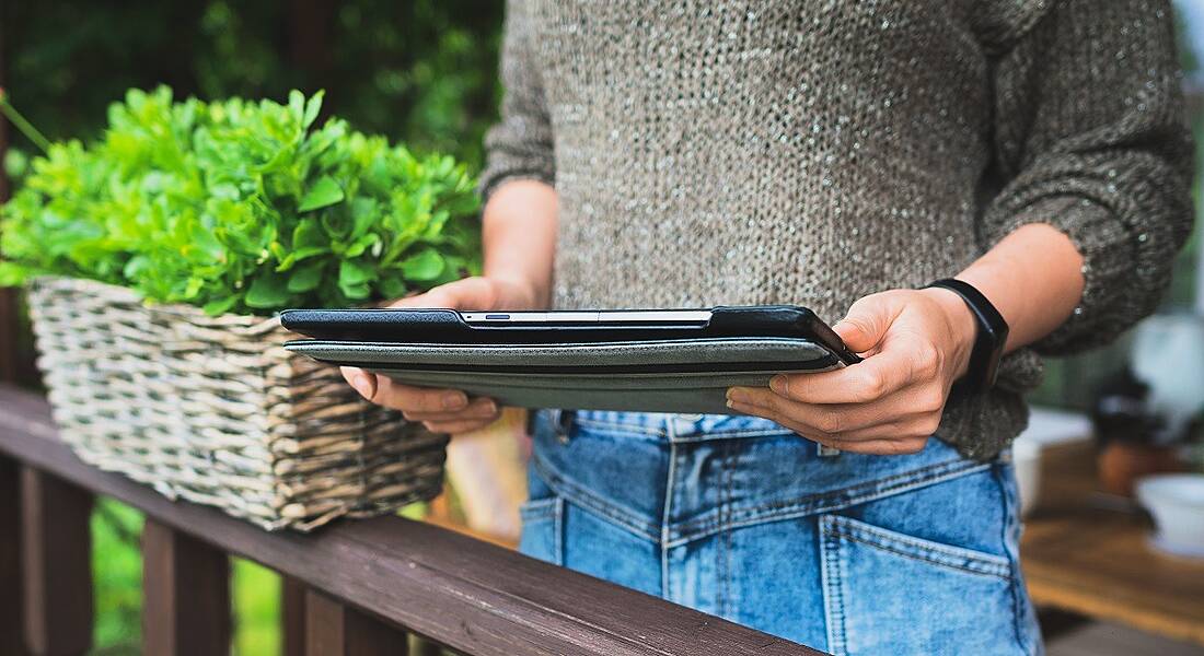 Woman in a light brown knitted jumper beside a flowerpot and holding a tablet device.