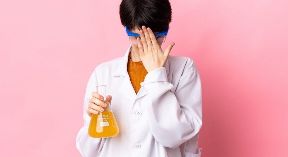 Woman in a white lab coat hiding her face and holding a beaker with a yellow liquid against a pink background.