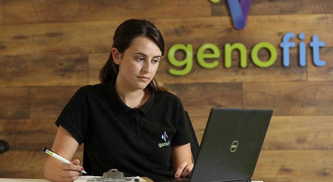 Research assistant Rachael Carr is sitting at a desk and working on a laptop in front of a sign reading Genofit on the wall behind her.