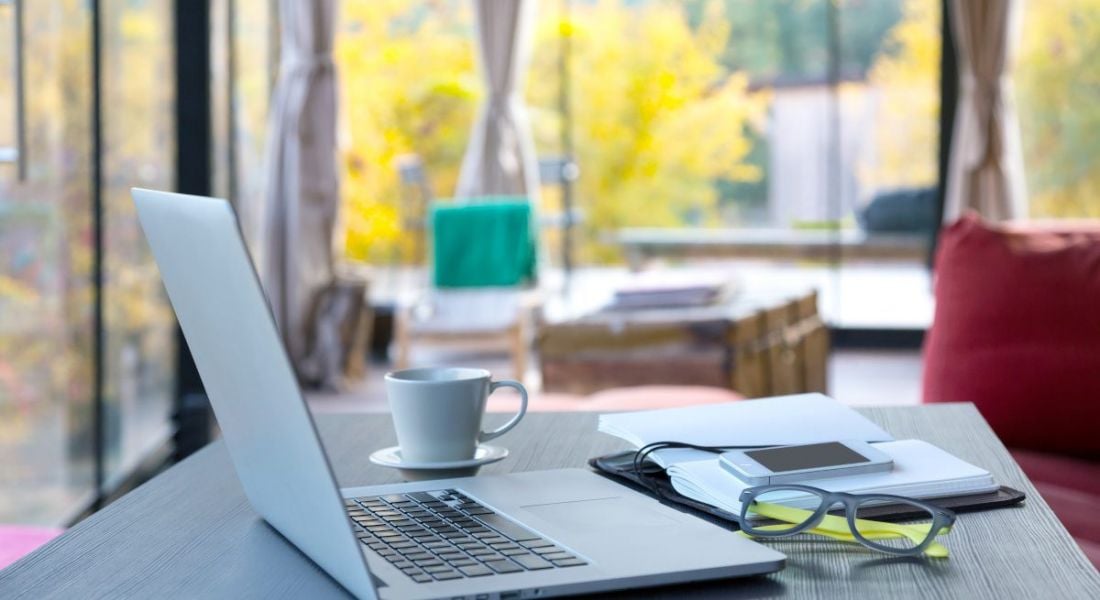 A laptop sits on a table with a notepad, a coffee cup and a pair of glasses. In the background, the room has large windows with colourful trees outside.