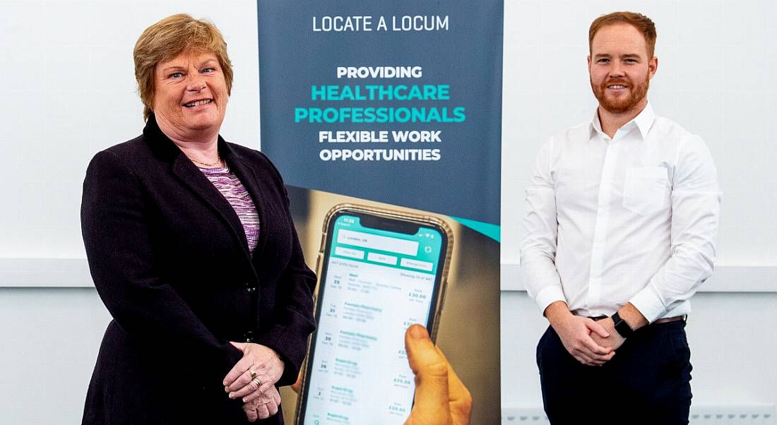 A woman and a man stand either side of a Locate a Locum pop-up banner, both standing with their hands clasped in front of their bodies.