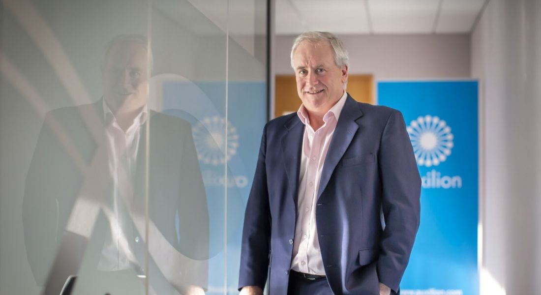 Auxilion CEO Philip Maguire is standing in the company’s offices and smiling into the camera while wearing a suit.