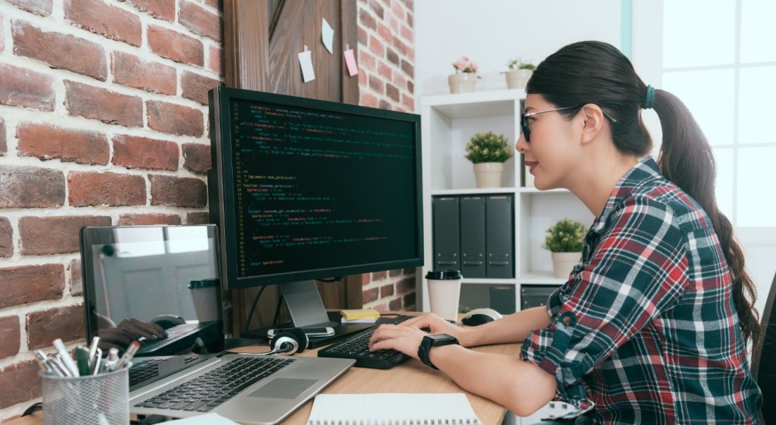 A young woman is sitting at a desk and working on coding on a computer.